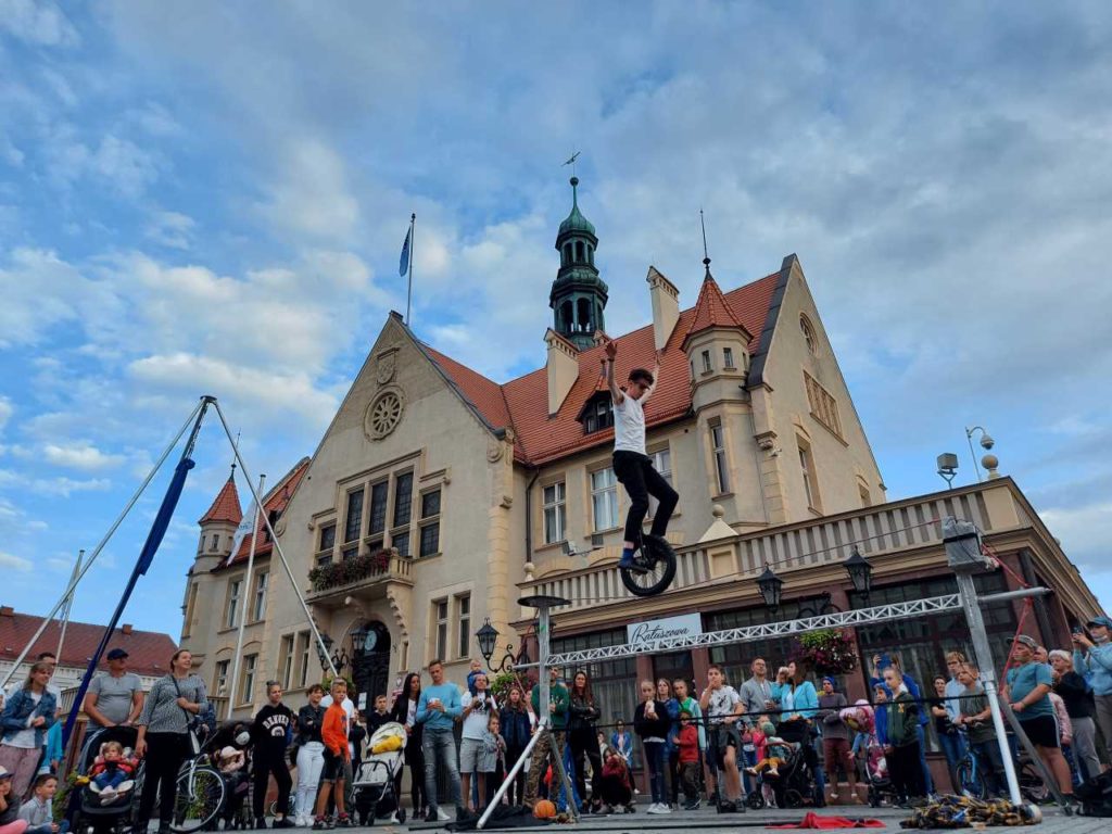 Street performer Sam Goodburn unicycling across the tightrope at BuskerBus 2021 in Krotoszyn. In the bak there is a townhall.