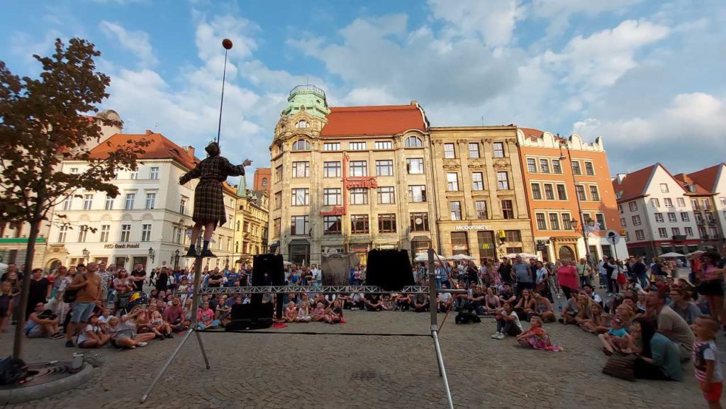 Tłum ogląda artystę ulicznego, który jest ubrany w szlafrok i stoi na metalowej konstrukcji i balansuje piłką znajdującą się na kiju.