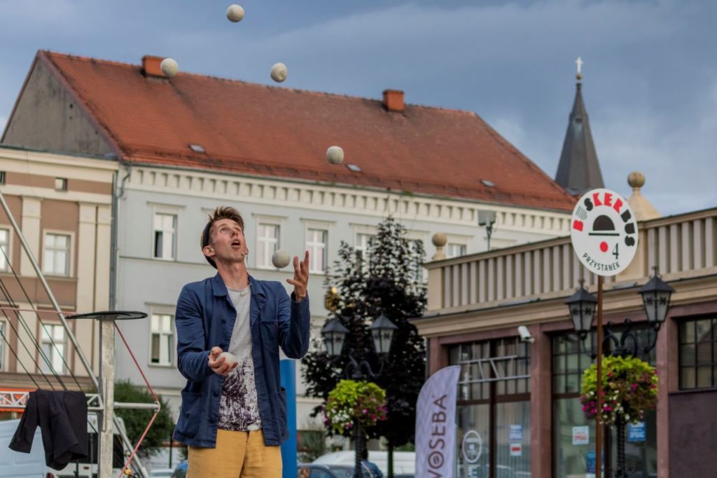 Busker Sam Goodburn żongluje sześcioma piłkami. Stoi obok znaku z logo festiwalu BuskerBus