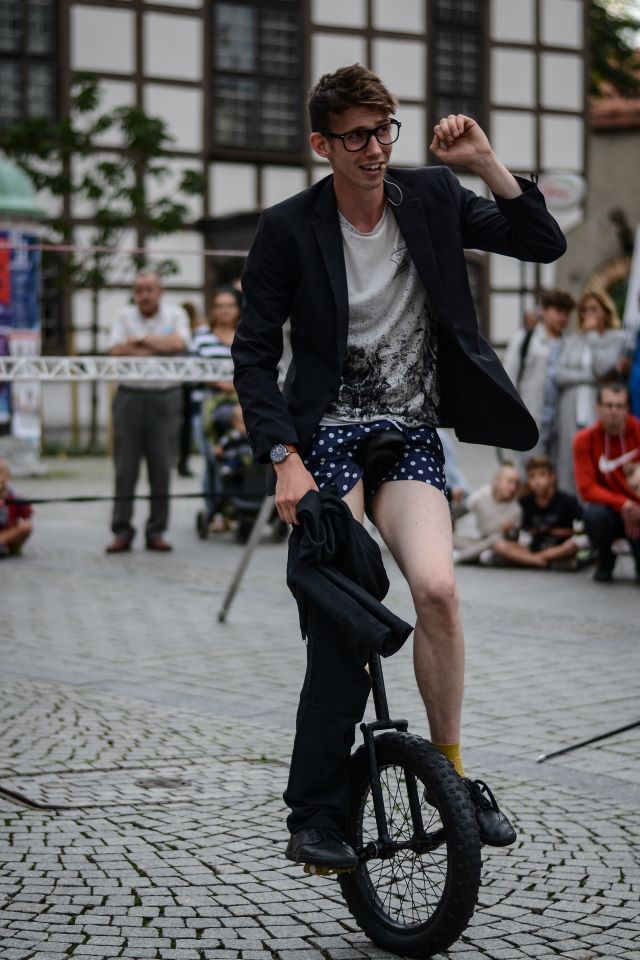 Artysta uliczny siedzi na monocyklu. Przytrzymuje spodnie, które ma założone tylko na jedną nogę.