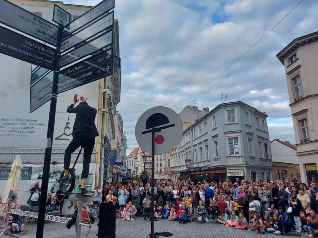 Tłum ogląda występ artysty ulicznego, który wsiada na monocykl znajdujący się na linie przymocowanej do metalowej konstrukcji.