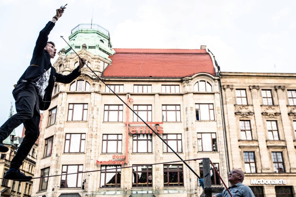 One man stands on a rope and tries to reach for a cookie that is attached to a fishing rod that the other man is holding.