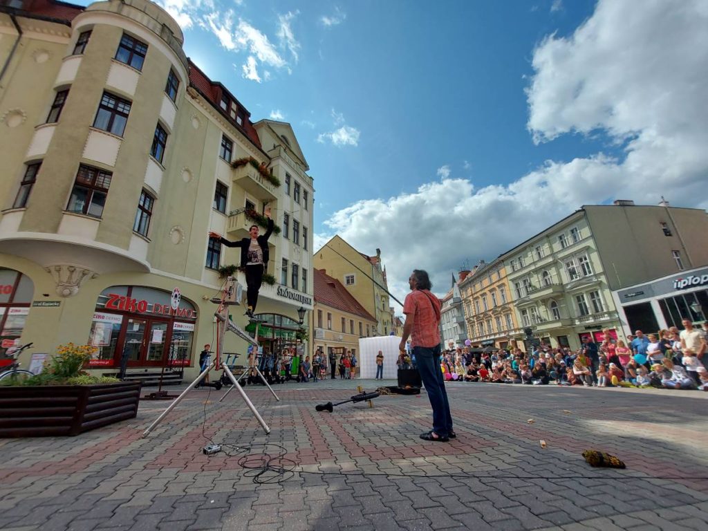 A man stands on a rope attached to a metal structure. He is fed a cookie that is attached to a fishing rod held by another man.