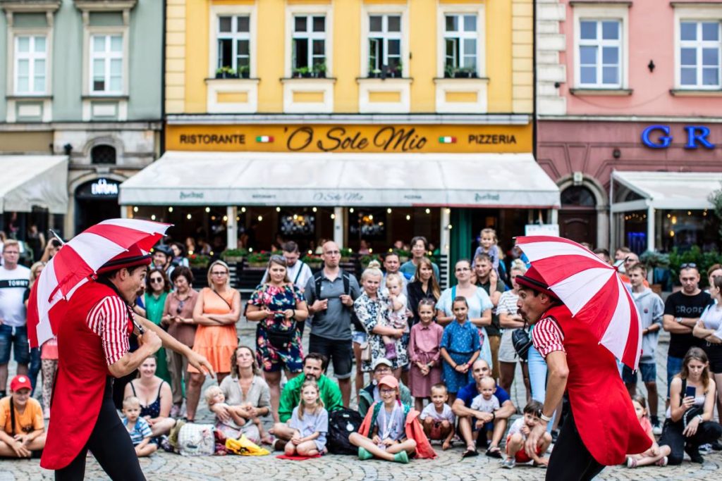 Identyczni bliźniacy prezentują publiczności uliczne przedstawienie cyrkowe. W rękach trzymają biało-czerwone parasolki.