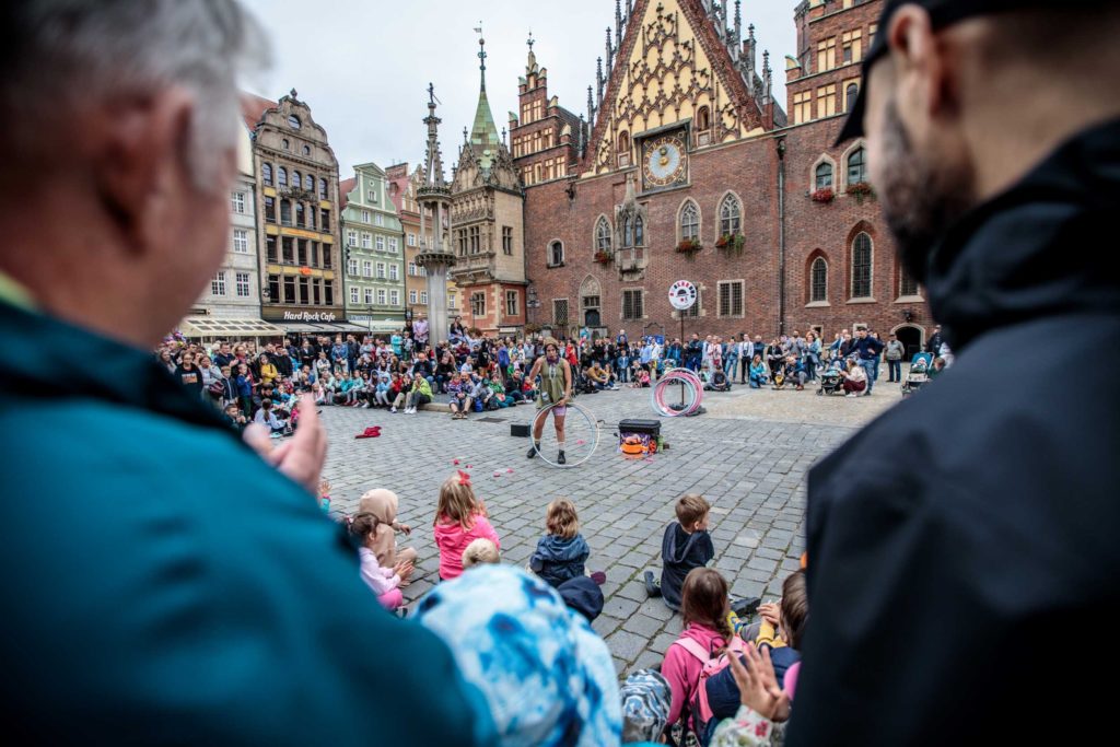 Tłum na festiwalu ulicznym ogląda pokaz artystki cyrkowej, która w ręce trzyma hula hop. W tle widać wrocławski ratusz.