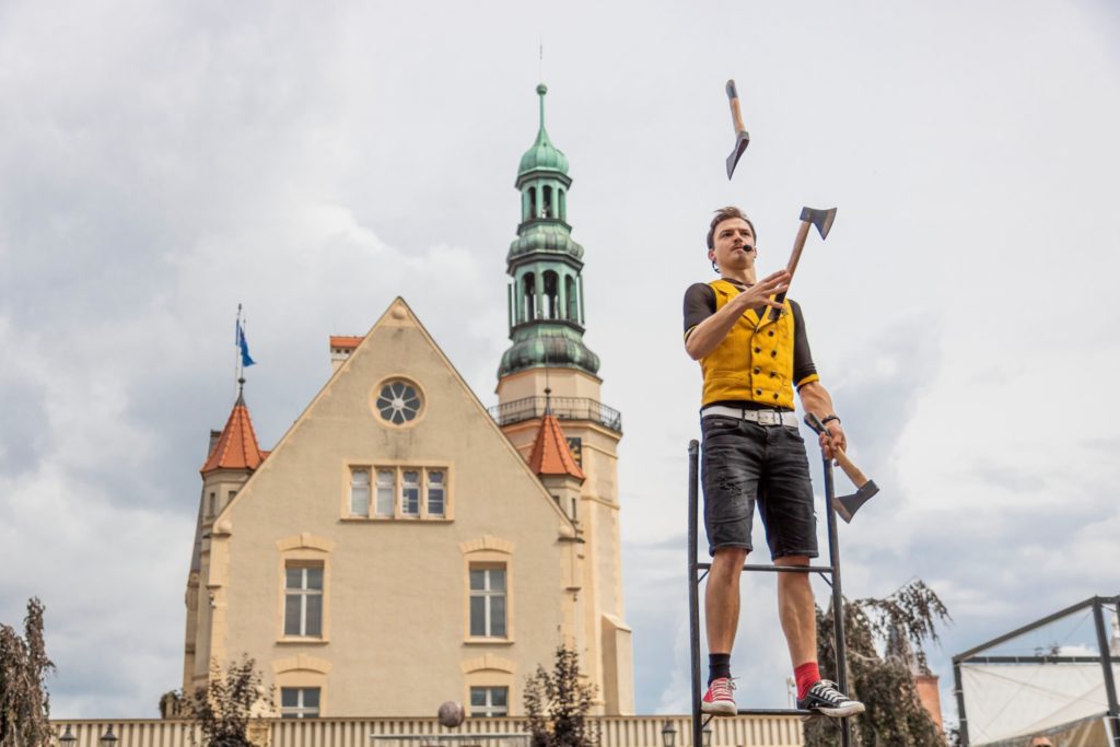 Krzysztof żongluje siekierami, balansując na drabinie. W tle krotoszyński Ratusz