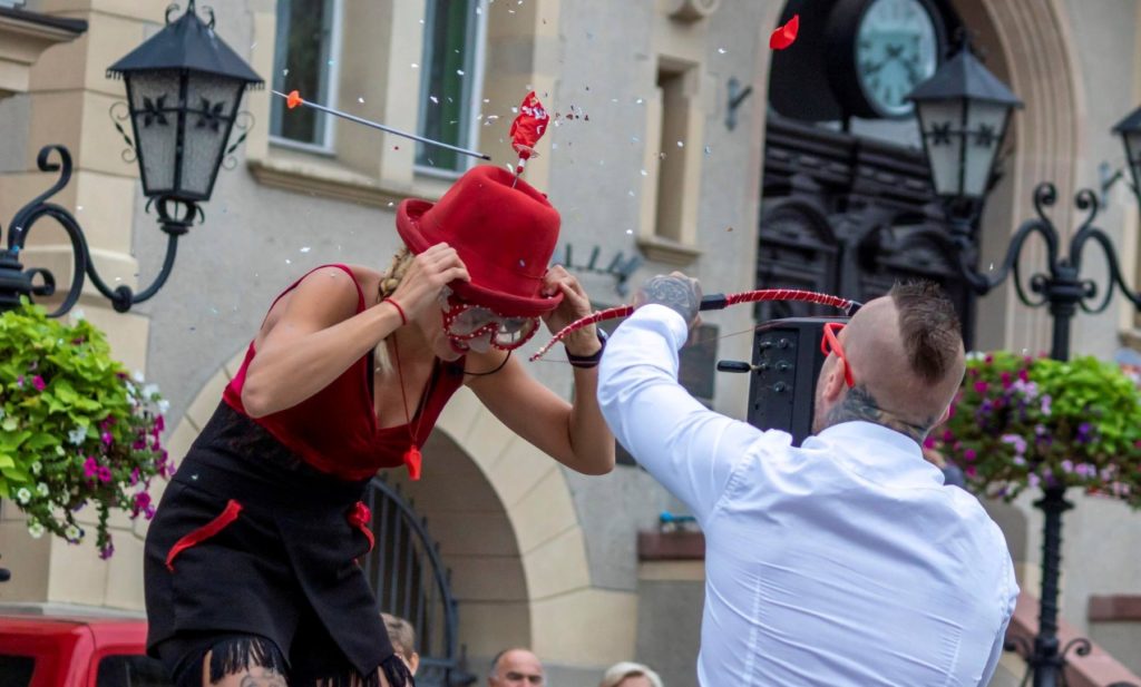 Mężczyzna strzela z łuku do balona przyczepionego do czerwonego kapelusza, który ma na głowie Gaia Ma