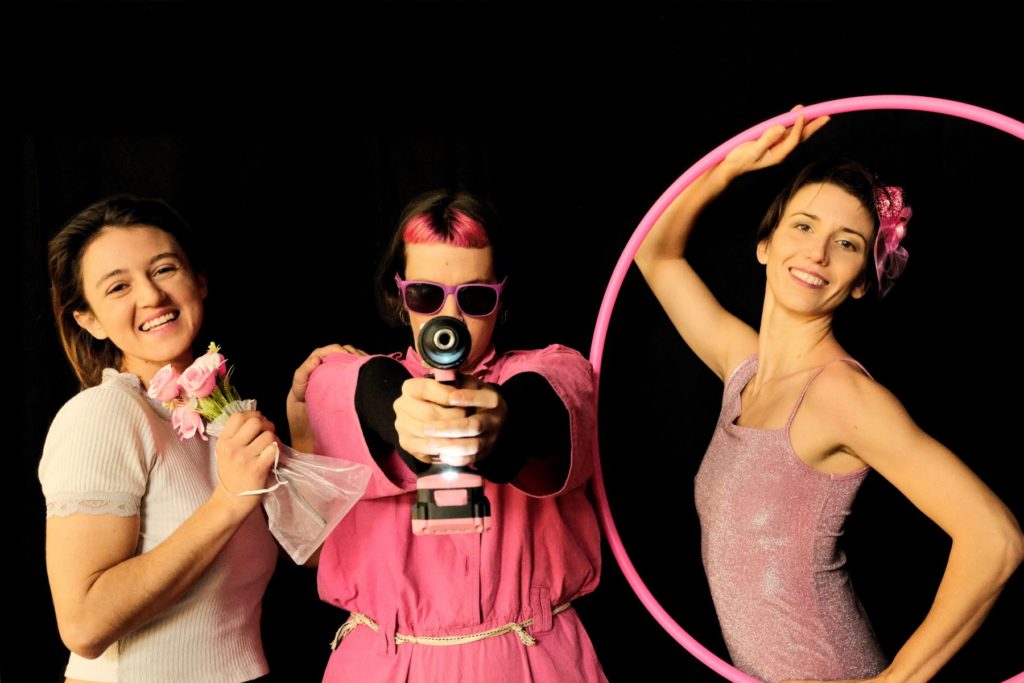Three women dressed in pink clothes. One holds flowers, the second one a drill and the third one hula hoop.