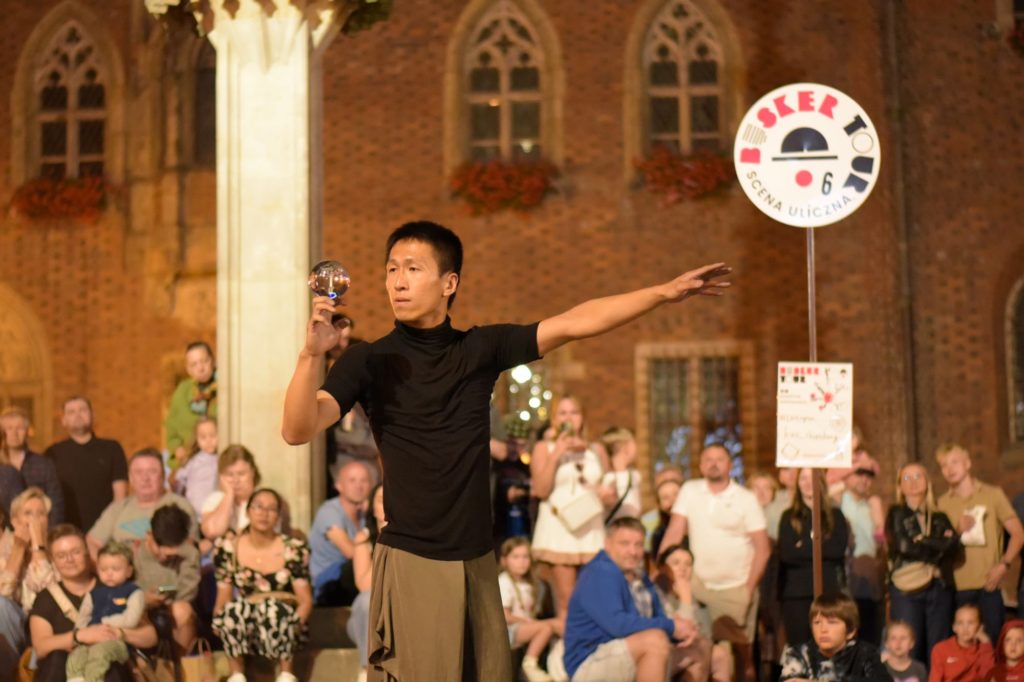 Busker Kuo Chien Hung występuje z kryształową kulą. W tle znak z logo festiwalu Busker Tour