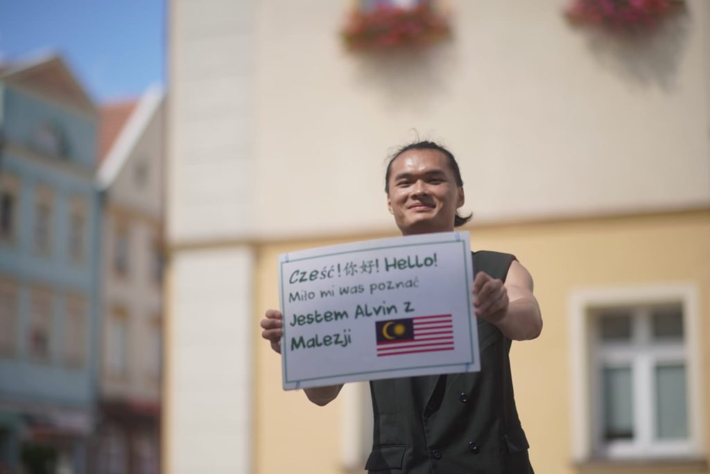 A street performer holds a poster with text in Polish hello nice to meet you. I'm Alvin from Malaysia