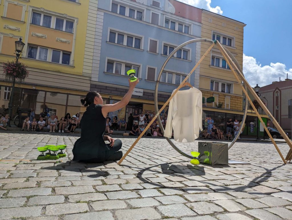 Artysta uliczny siedzi na ziemi. Obok rekwizyty cyrkowe: koło Cyra i diabolo