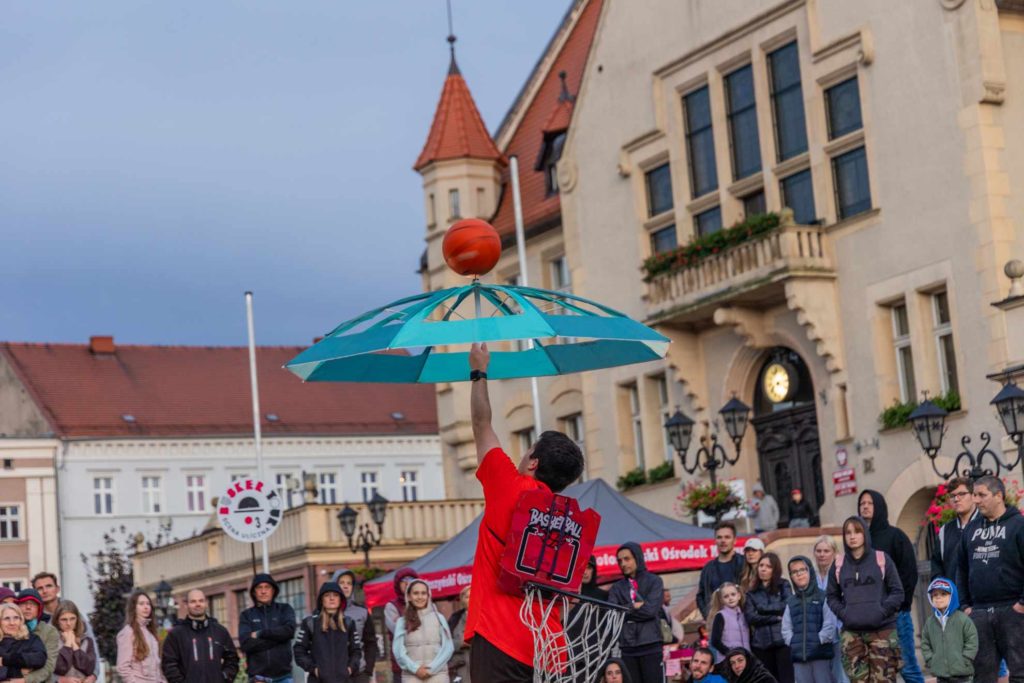 Artysta uliczny Stefan Florescu, trzyma niebieski parasol, na którego czubku kręci się piłka do koszykówki. W tle publiczność, ratusz  w Krotoszynie i znak festiwalu Busker Tour