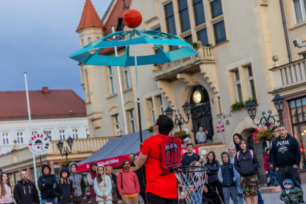 Street performer Stefan Florescu holds an umbrella with a basketball spinning on top of it. In the background, there is a sign with the Busker Tour festival logo and the town hall in Krotoszyn.