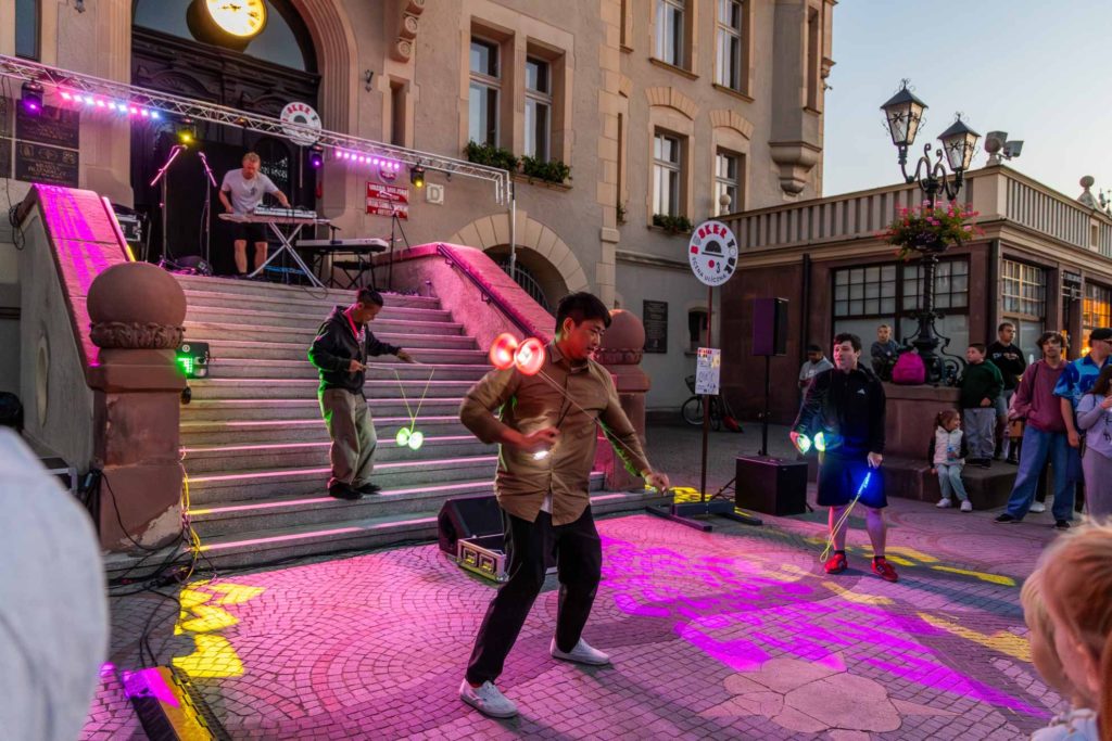 Buskers performing a group show at Busker Tour. Three artists performing tricks with diabolo, with a musician visible on the stairs in the background.