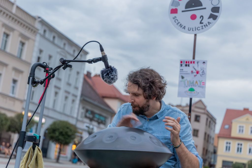 Artysta uliczny TIMAY gra na hanpanie. W tle znak festiwalu Busker Tour